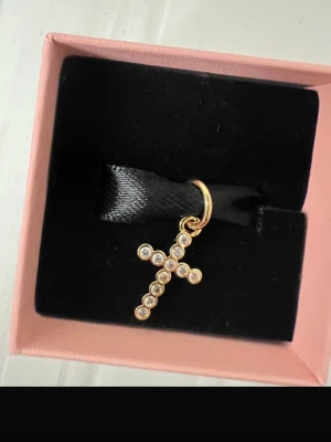 Pandora korsberlock i guld med stenar - Säljer en snygg korsberlock från Pandora i guldton med glittrande stenar. Perfekt att hänga på ett armband eller halsband för att ge din look lite extra bling. Kommer i original ask från Pandora.