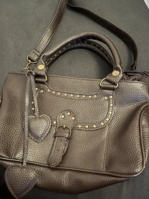 Brun handväska med nitar och hjärtan - Säljer en brun handväska i skinn med snygga guldfärgade nitar och dekorativt bälte framtill. Väskan har både handtag och axelrem samt två hjärtformade detaljer som hänger på framsidan. Perfekt för dig som gillar unika accessoarer!