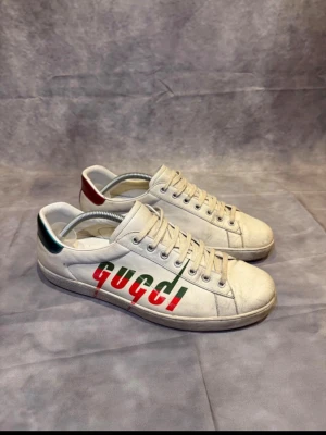 Gucci sneakers med logga och färgdetaljer - Snygga vita sneakers från Gucci med stor logga i rött och grönt på sidan. Skorna har snörning och platt sula, samt metallic-detaljer i rött och grönt på hälen. Tillverkade i skinn och har en klassisk rund tå. Perfekta för dig som gillar exklusiv streetstyle.