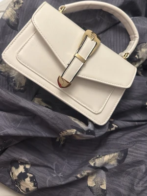 Vit handväska med gulddetaljer - Snygg vit handväska som är helt ny,  den har skinnimitation med stilren design och guldfärgat spänne framtill. Väskan har ett praktiskt handtag, en ficka på baksidan och dragkedja inuti för extra förvaring. Perfekt storlek för det viktigaste och passar till många olika outfits.