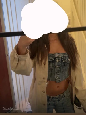 Blå croppad denim topp - Säljer en blå croppad-topp i denim med snygga silverfärgade knappar framtill. Toppen har en cool, edgy look med råa kanter och är axelbandslös. Perfekt för dig som vill sticka ut med en trendig jeansstil.