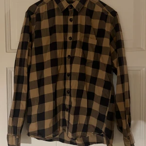 Rutig beige och svart skjorta - Säljer en långärmad skjorta med stort rutmönster i beige och svart. Skjortan har klassisk krage, bröstficka och knappar framtill. 100% bomull. Slitage bakom nedre knappen, syns ej när knappen är stängd. Se bild.