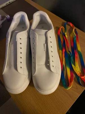 Alexander McQueen vita sneakers med regnbågsdetaljer - Säljer ett par vita Alexander McQueen sneakers i skinn med regnbågsfärgad logga på hälen och tillhörande färgglada skosnören. Klassisk rund tå och platt sula, riktigt snygga och unika detaljer som sticker ut. Perfekt för dig som gillar färg och stil.