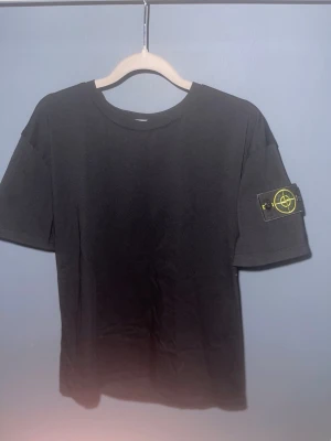 Svart Stone Island t-shirt - Säljer en svart t-shirt från Stone Island med klassisk rund hals och korta ärmar. På vänster ärm sitter den ikoniska Stone Island patchen med gul brodering och knappar. T-shirten är gjord i mjuk bomull och har en clean, stilren look.