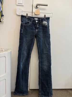 Blå bootcut jeans med broderade fickor - Snygga mörkblå jeans med bootcut-snitt och kontrastsömmar. Baktill finns två fickor med detaljerad brodering och paljetter i beige och silver som ger extra stil. Jeansen har klassisk femficksdesign och är gjorda i jeansmaterial med en skön passform. Köptes för 400kr