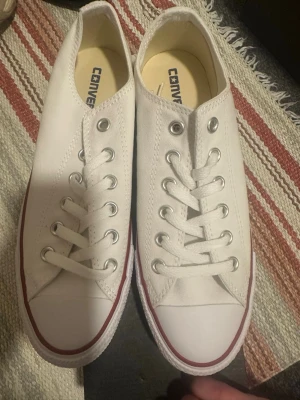 Vita Converse All Star sneakers - Säljer ett par klassiska vita Converse All Star sneakers i storlek 39.5. Skorna har lågt skaft, vit canvas, snörning med metallhål och en vit gummisula med röd rand. Perfekta för dig som gillar enkel och tidlös stil.