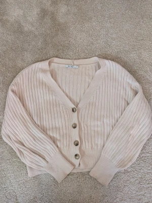 Beige ribbad kofta från NA-KD - Superfin beige kofta från NA-KD i ribbstickad bomull. Modellen är croppad med v-ringning, stora knappar framtill och långa ärmar med breda muddar. Perfekt att slänga över en topp för en chill look.