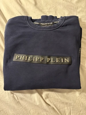 Mörkblå sweatshirt Philipp Plein - Säljer en mörkblå sweatshirt från Philipp Plein med stort logotyptryck i silver på bröstet. Tröjan har rund halsringning, långa ärmar och ribbade muddar. Tillverkad i mjuk bomull för skön känsla och stilren look.