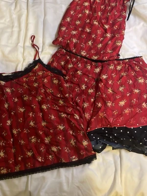 Röd blommig linnetopp från ottod'Ame - Säljer en röd linnetopp från ottod'Ame med blommigt mönster och svarta vertikala ränder. Toppen har tunna axelband och är dekorerad med svart spets längs halsringningen och nederkanten. Materialet är mjukt och glansigt, perfekt för sommarens varma dagar.