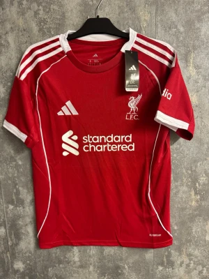 Liverpool Isak #9 fotbollströja - Ny storlek S - Säljer en röd Liverpool FC matchtröja från Adidas med vita detaljer och tryck. Tröjan har klubbmärke, sponsorlogga och nummer 9 med namnet Isak på ryggen. Klassisk design med vita ränder på axlarna och korta ärmar. Perfekt för fotbollsfans!