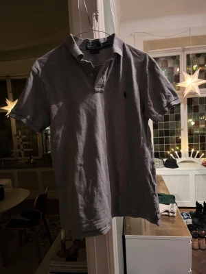 Grå pikétröja från Polo Ralph Lauren - Säljer en grå pikétröja från Polo Ralph Lauren i slim fit-modell. Tröjan har korta ärmar, klassisk krage och två knappar framtill. Materialet är mjuk bomull och den har den ikoniska broderade loggan på bröstet.