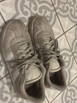 Beige Adidas sneakers med mocka - Säljer ett par beige Adidas sneakers med klassiska vita ränder på sidan. Skorna har rund tå, platt sula och snörning. Ovandelen är i mocka och insidan är fodrad med textil. Perfekt för dig som gillar stilrena och neutrala färger. De är väldigt smutsiga, vissa fläckar går nog bort men de svarta fläckarna vid ränderna går inte bort så lätt. 