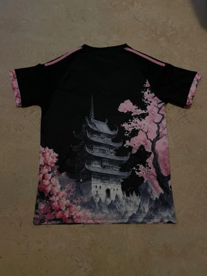 Japan Adidas fotbollströja  - Unik svart fotbollströja från Adidas med Japan-tema, prydd med en stor drake, körsbärsblommor och ett pagodmotiv. Rosa detaljer på axlar och ärmslut, samt v-ringad hals. Perfekt för dig som gillar coola prints och asiatiska influenser.
