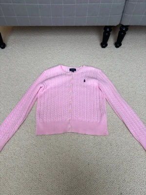 Rosa kabelstickad kofta Polo Ralph Lauren - Supersöt rosa kabelstickad kofta från Polo Ralph Lauren med klassisk logga broderad på bröstet. Koftan har rund hals, långa ärmar och knäppning framtill med vita knappar. 