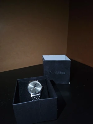 Klocka Axcent - Silverfärgad klocka från Axcent med metallarmband och datumvisning. Den är helt ny och extra band för förlängning inkluderas. Lådan kan stängas fint och detta kan användas som en bra present. Inget är skadat! Den här klockan är för de som letar efter något lyxigt och äkta!