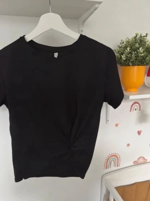 Svart omlott topp med twist - Säljer en svart topp med korta ärmar och snygg twist-detalj framtill. Toppen har rund hals och är tillverkad i mjukt material som känns skönt mot huden. Perfekt att matcha med jeans eller kjol för en cool look.