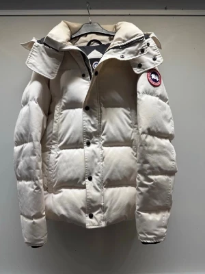 Vit pufferjacka från Canada Goose - Säljer en snygg vit pufferjacka från Canada Goose med ikonisk patch på ärmen. Jackan har hög krage, avtagbar huva och knappar samt dragkedja framtill. Fylld med dun och har en clean, boxig look som är perfekt för kalla dagar.