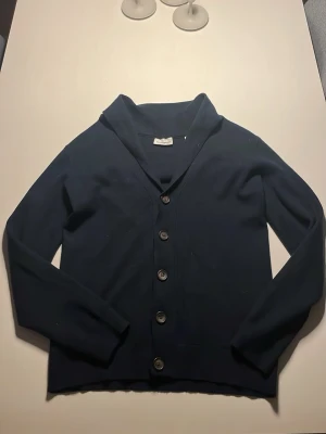 Mörkblå cardigan från Lindbergh - Snygg mörkblå cardigan från Lindbergh med klassisk v-ringning och fem stora knappar framtill. Cardiganen har långa ärmar och är tillverkad i mjuk bomull, perfekt för lager-på-lager under kyliga dagar. Enkel och stilren design som funkar till många outfits. Storlek S men passar också på M. 🔥OBS FAST PRIS🔥