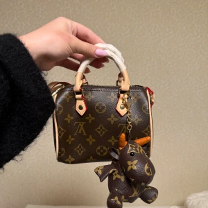 Monogram handväska - Säljer en liten handväska med klassiskt monogrammönster i brunt och beige. Väskan har ljusa handtag, guldfärgade detaljer och dragkedja. Medföljer en söt nyckelring. Perfekt accessoar för att lyfta din look.