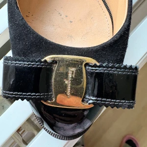 Svarta pumps från Salvatore Ferragamo - Stilrena svarta pumps från Salvatore Ferragamo i mocka och lackat skinn. Skorna har en dekorativ rosett framtill med guldfärgad metallplatta och rund tå. Klacken är bred och stabil, vilket ger både höjd och komfort. Perfekta för dig som gillar klassisk och lyxig stil.