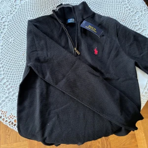Svart half zip tröja från Polo Ralph Lauren - Svart långärmad tröja från Polo Ralph Lauren med half zip och röd broderad logga på bröstet. Tröjan har en klassisk krage med vit rand och är tillverkad i ett strukturerat bomullstyg. Perfekt för en stilren och avslappnad look.