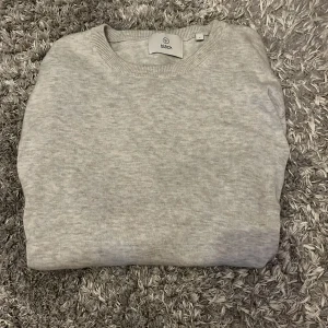 Ljusgrå stickad tröja från Bläck - En stilren ljusgrå stickad tröja från Bläck i storlek S. Tröjan har rund halsringning, ribbade muddar och är långärmad. Perfekt för lager-på-lager och enkel att matcha med jeans eller chinos. Nypris 600