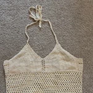 Beige virkad linnetopp med knyt - Säljer en beige virkad linnetopp med smala axelband och knyt i nacken. Toppen har en luftig, mönstrad virkning och v-ringad hals. Perfekt för varma dagar och ger en bohemisk vibe.