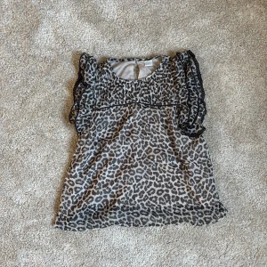 Leopardmönstrad blus från Lindex - Säljer en snygg blus från Lindex med grått och svart leopardmönster. Blusen har volangdetaljer på ärmarna och en rund halsringning med knapp i nacken. Tillverkad i ett lätt och luftigt material.
