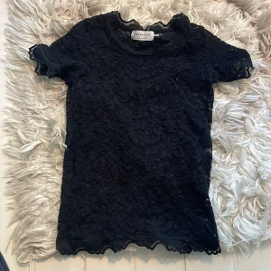 Svart spetsblus från Rosemunde - Supersnygg svart blus från Rosemunde med korta ärmar och genombruten spets med blommigt mönster. Blusen har vågig kant vid hals och ärmslut, och är perfekt för dig som gillar romantiska detaljer och en feminin look.