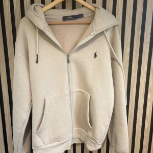 Beige hoodie från Polo Ralph Lauren - Beige hoodie från Polo Ralph Lauren med dragkedja, huva och snörning. Klassisk broderad logga på bröstet och stora fickor framtill. Mjuk insida och relaxed fit, perfekt för chill dagar. EJ EKTA!