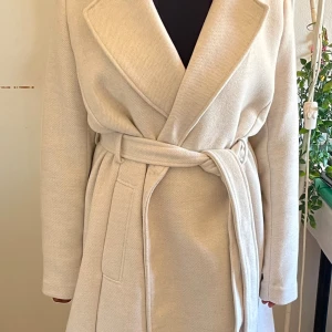 Beige kappa från H&M med bälte - Stilren beige kappa från H&M i storlek 44. Kappan har breda slag, knyts med bälte i midjan och har diskreta fickor på sidorna. Lång modell med slits bak och klassisk look, perfekt för dig som gillar minimalistisk stil. Kappa, storlek 44 (EU motsvarar ca L) Använd lite grann men i fint skick.Bilden visar kappan på en person som är ca 165 cm lång🧍‍♀️