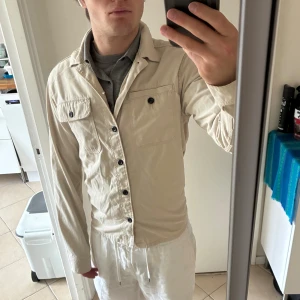 Beige overshirt från Jack & Jones XS - Snygg beige overshirt från Jack & Jones Core med svarta knappar och två bröstfickor. Skjortan har klassisk krage och är tillverkad i mjuk bomull. Perfekt lager-på-lager-plagg med stilren look och diskret logga på ärmen. Bra skick 