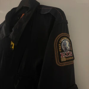 Svart vindjacka Parajumpers XX Large - Svart vindjacka från Parajumpers med ikonisk patch på ärmen och flera praktiska fickor framtill. Jackan har orange detaljer, klassisk krage och stängs med både dragkedja och knappar. Slim fit-modell, perfekt för dig som gillar stilren och funktionell design.