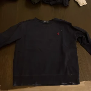 Mörkblå tröja från Polo Ralph Lauren - Säljer en klassisk mörkblå tröja från Polo Ralph Lauren med rund hals och lång ärm. Tröjan har den ikoniska röda loggan broderad på bröstet och är tillverkad i mjuk bomull. Perfekt för en stilren och avslappnad look.