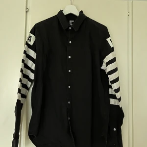 Svart skjorta från Hood By Air - Svart skjorta från Hood By Air med vita ränder på ärmarna och stora HBA-tryck på ryggen och axlarna. Skjortan har knappar framtill och är tillverkad i 100% bomull. Kan mötas upp på Södermalm. Storlek L men skulle säga sitter mer som en M.