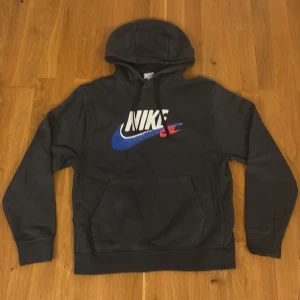 Grå Nike hoodie med färgglad logga - Grå hoodie från Nike i 100% bomull med stor vit, blå och röd Nike-logga framtill och mindre loggor i samma färger på ryggen. Klassisk känguruficka, huva med snörning och mjuk insida. Perfekt för dig som gillar sportig och street-inspirerad stil. Pris går att diskutera🙌