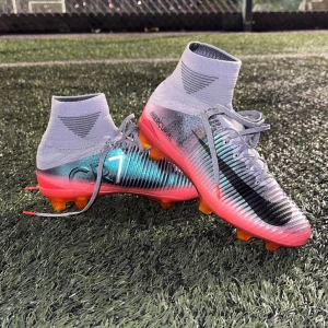 Nike Mercurial CR7 fotbollsskor - Säljer ett par Nike Mercurial CR7 fotbollsskor med högt skaft och snörning. Skorna har en snygg färgskiftning i silver, grått och rosa med svarta detaljer och orange dobbar. Perfekta för dig som vill sticka ut på planen och få grymt grepp på konstgräs.