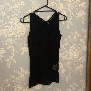 Svart spetsig ärmlös topp från AMISU - Svart ärmlös topp från AMISU i storlek XS. Toppen är helt i spets med ett genomskinligt, nätliknande mönster och rund halsringning. Perfekt för att skapa en edgy och trendig look, funkar snyggt över ett linne eller bralette.