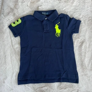 Marinblå pikétröja från Ralph Lauren - Snygg marinblå pikétröja från Ralph Lauren med klassisk krage och korta ärmar. Stor gul logga på bröstet och siffra på ärmen i samma färg. Tillverkad i bomull, perfekt för en sportig och stilren look.