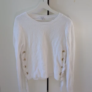 Vit stickad tröja med snörning från Amisu - Vit stickad tröja från Amisu med snygga snörningsdetaljer i sidorna och guldiga öljetter. Tröjan har rund halsringning och långa ärmar, perfekt för lager-på-lager under hösten eller vintern.