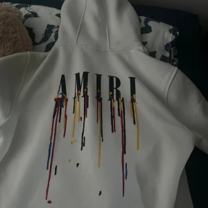 Vit hoodie från Amiri med färgstänk - Vit hoodie från Amiri med stor huva och coolt tryck på bröstet där AMIRI-loggan droppar i färger som röd, gul, blå och svart. Hoodien har metallöljetter vid huvan och är i mjukt material, perfekt för dig som gillar streetwear med edge.