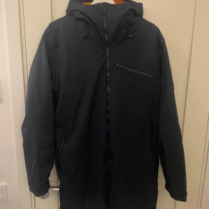 Mörkblå parkas från Peak Performance - Snygg mörkblå parkas från Peak Performance med huva och flera dragkedjeförsedda fickor. Jackan har en stilren design med logotyp-patch på ärmen och är tillverkad i slitstarkt syntetmaterial. Perfekt för kalla dagar och har en längre passform.