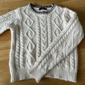 Vit stickad tröja från Mango - Säljer en vit stickad tröja från Mango med grovt flätmönster och ribbade muddar. Rund halsringning med svartvit detalj som ger en snygg kontrast. Perfekt för kyliga dagar och enkel att matcha med jeans eller kjol.