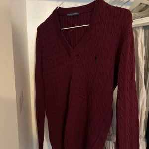 Vinröd kabelstickad tröja Ralph Lauren - Säljer en vinröd kabelstickad tröja från Ralph Lauren med v-ringning och  broderad logga på bröstet. Tröjan har lång ärm och klassisk passform, perfekt för lager-på-lager under hösten och vintern .Strleken på den är L men passar även bra för M 