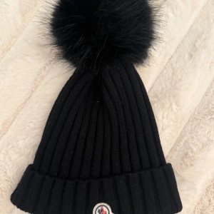 Svart Moncler mössa med pälsboll - Snygg svart ribbstickad mössa från Moncler med stor fluffig pälsboll på toppen och klassisk Moncler-logga framtill. Perfekt accessoar för kalla dagar, både stilren och trendig. Tillverkad i mjukt material som håller dig varm hela vintern.