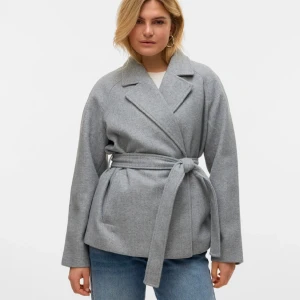 Grå omlottjacka med bälte - Snygg grå jacka i omlottmodell med bälte i midjan och klassisk krage. Jackan har lång ärm och en stilren, rak passform. Perfekt för dig som gillar en clean och enkel look.