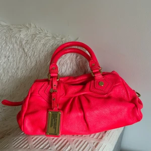 Marc by Marc Jacobs rosa handväska - Cool handväska från Marc by Marc Jacobs i knallig rosa färg med guldiga metalldetaljer och avtagbar axelrem. Väskan är gjord i skinn och har ett snyggt innerfoder med svartvitt mönster och dragkedja upptill. Perfekt för dig som vill sticka ut med din look.