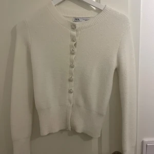Vit kofta med pärlknappar från Zara - Superfin vit kofta från Zara med långa ärmar och rund hals. Koftan har dekorativa pärlknappar framtill och är stickad i ett mjukt material. Väldigt bra skick, säljer då den är för liten.