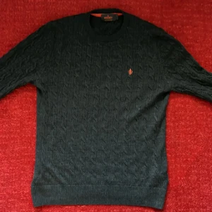 Mörkgrå kabelstickad tröja Morris - Kabelstickad mörkgrå tröja från Morris i 100% merinoull. Klassisk rund halsringning och diskret orange logga broderad på bröstet. Perfekt för dig som gillar stilrena och tidlösa plagg med premiumkänsla.