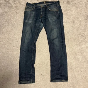 Mörkblå jeans från Lee Darren - Säljer ett par klassiska mörkblå jeans från Lee med raka ben och snygga slitningar. Jeansen har fem fickor, gylf med dragkedja och Lee-logga på bakfickan. Perfekta för dig som gillar en tidlös och enkel stil.